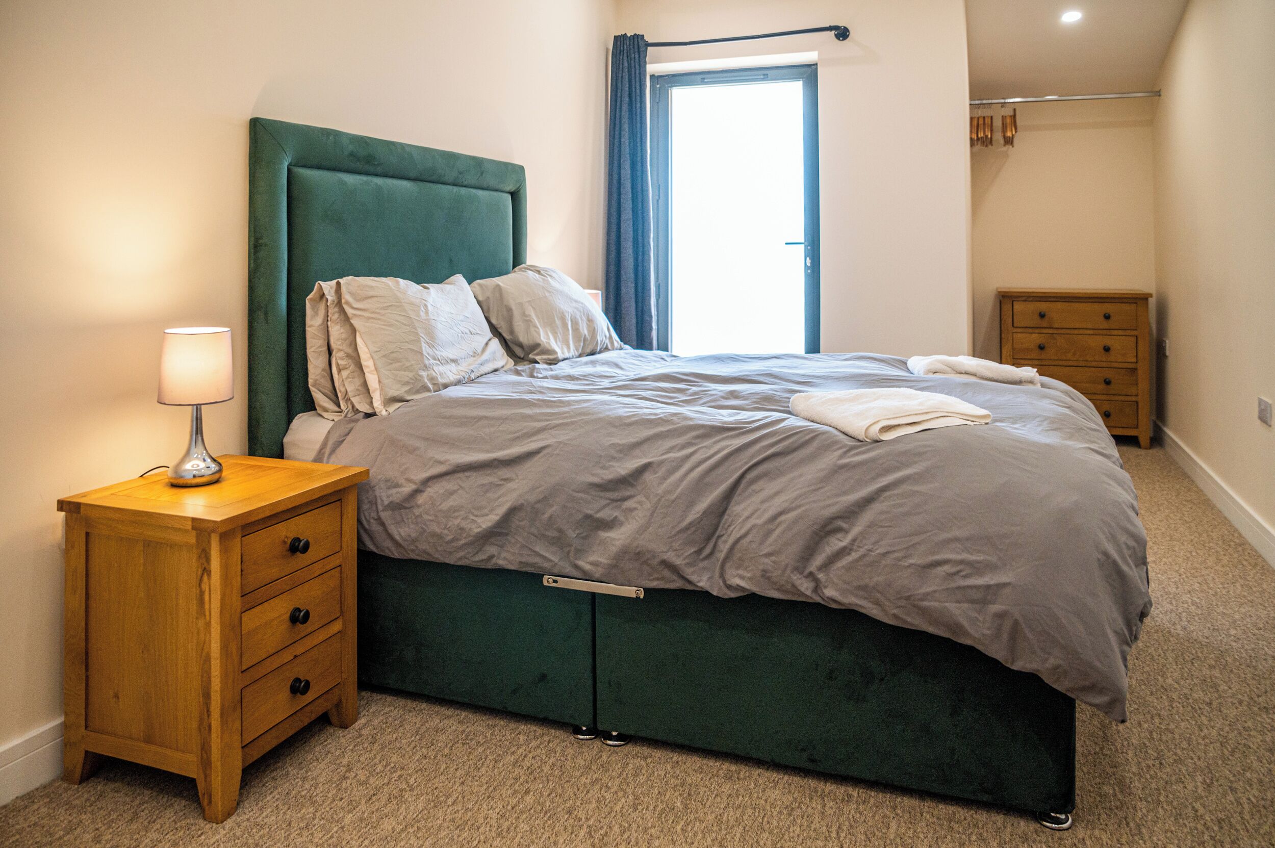 A neatly made bed with a green headboard, a wooden nightstand with drawers, and a lamp.
