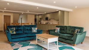 A living room with two green sofas, a wooden coffee table, and a kitchen in the background. - Bears Court Barn 6 (Cheltenham)