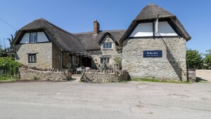 A traditional stone building with a thatched roof, a sign indicating it is open all day, and a stone wall in front. - Hazel (Witney)