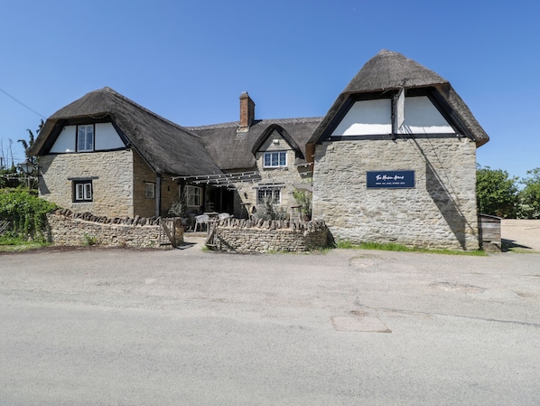 A traditional stone building with a thatched roof, a sign indicating it is open all day, and a stone wall in front. - Hazel (Witney)
