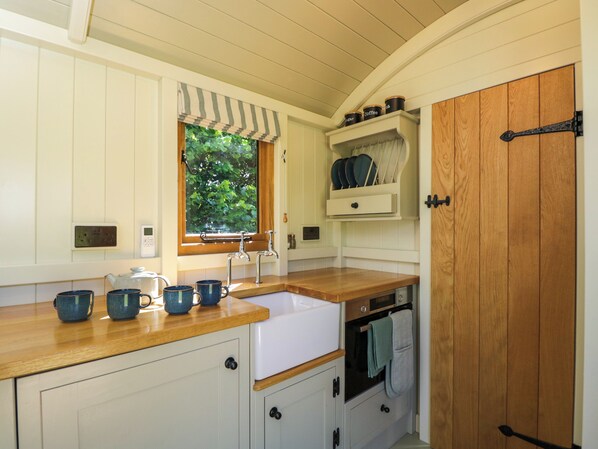A compact kitchen with a wooden countertop, white cabinetry, a window with a view of greenery, a built-in oven, and a wooden door. - Hazel (Witney)
