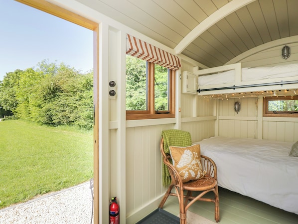 A cozy bedroom with a bunk bed, a wicker chair with a pillow, a window with a striped valance, and a fire extinguisher.