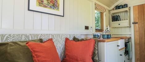 A cozy room with a patterned sofa, a framed floral artwork, a kitchen area with cabinets and utensils, and a window with curtains.