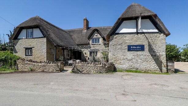 A traditional stone building with a thatched roof, a sign indicating it's open all day, and a stone wall in front.