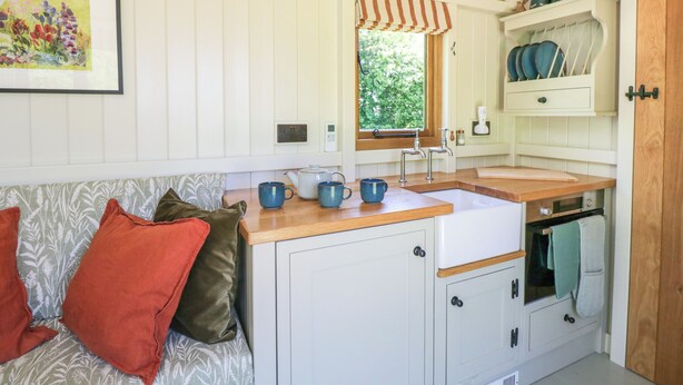 A compact kitchen with white cabinets, a wooden countertop, and a window with a striped awning. - Blackthorn (Witney)