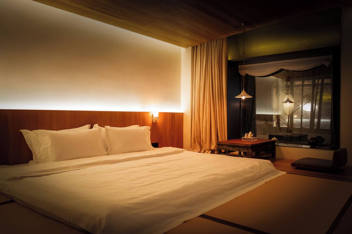 A neatly made bed in a room with a wooden headboard, a nightstand with a lamp, and a view of trees through a window with sheer curtains.