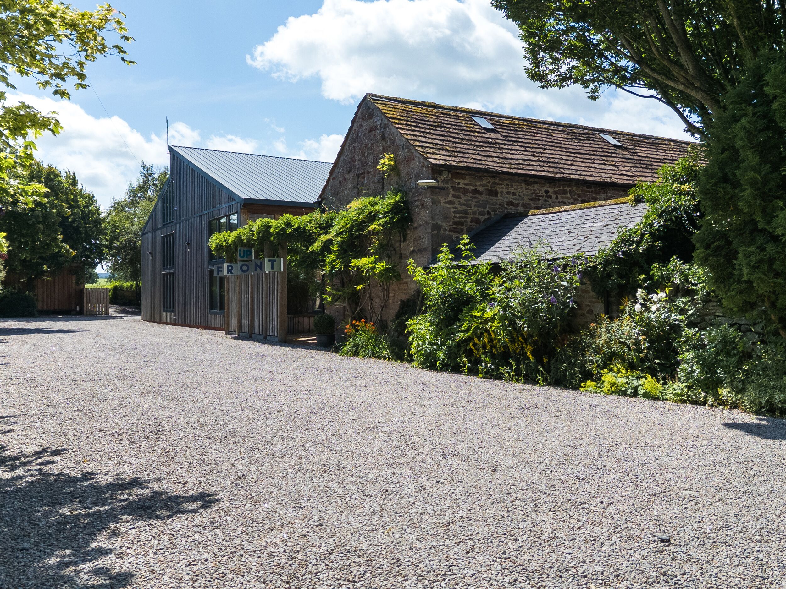 A gravel driveway leading to a stone building with a sloped roof, a modern metal structure, and lush greenery.
