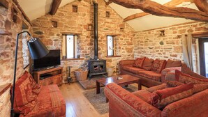 Ein gemütliches Zimmer mit Steinwand, Holzbalken, Kamin, einer Couch und einem Fernseher.
