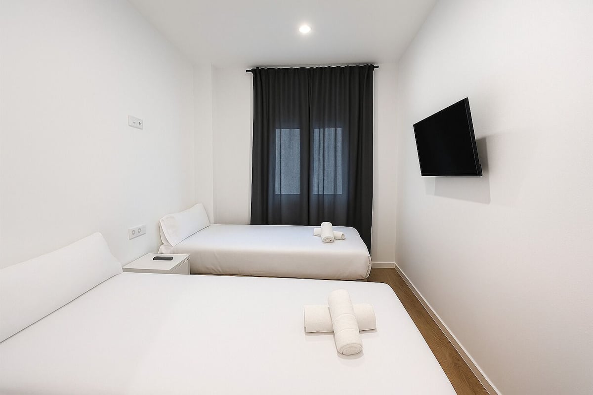Una habitación de hotel con dos camas individuales, un televisor de pantalla plana montado en la pared y una ventana con cortinas oscuras.