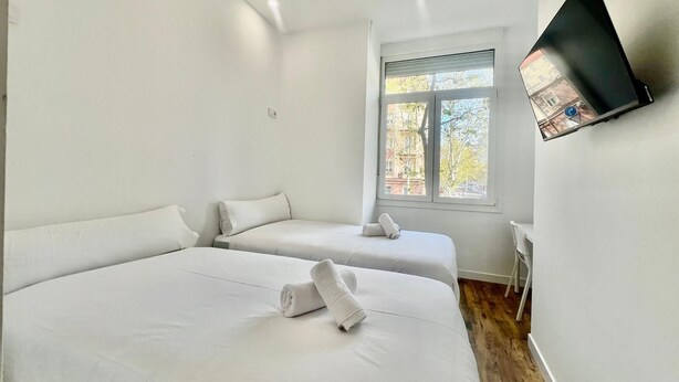 A modern hotel room with two beds, a TV mounted on the wall, and a window with a view of buildings and trees. - Hostal Bernabeu (Madrid)