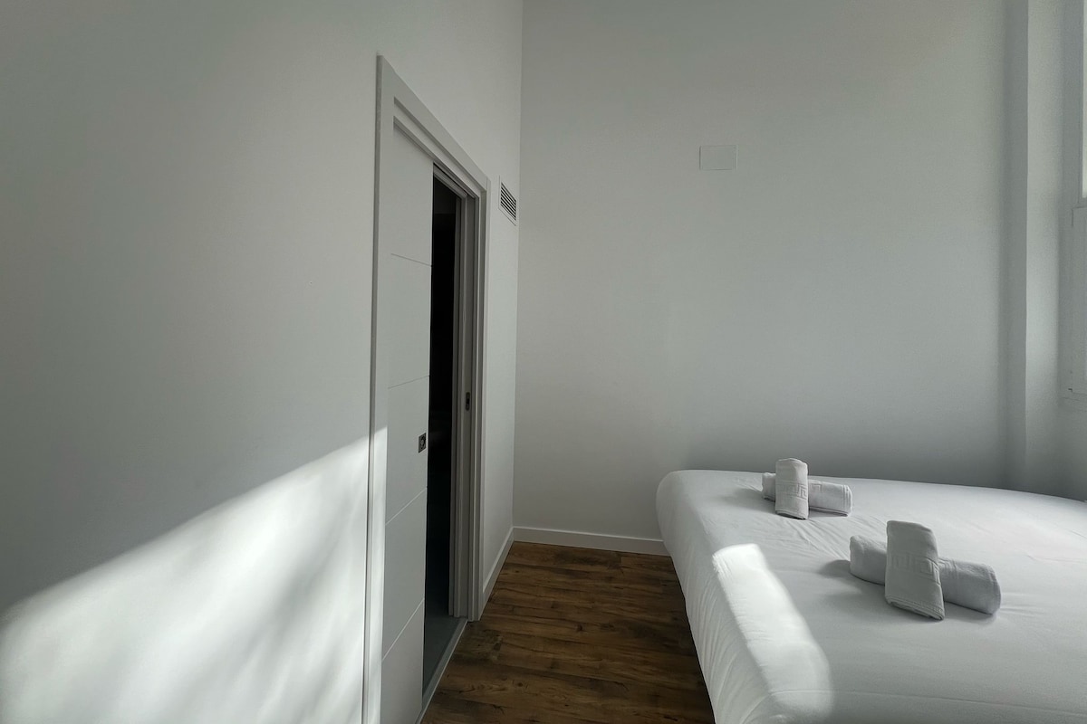 A hotel room with a wooden floor, a bed with white linens and towels, and a white door with a small vent.
