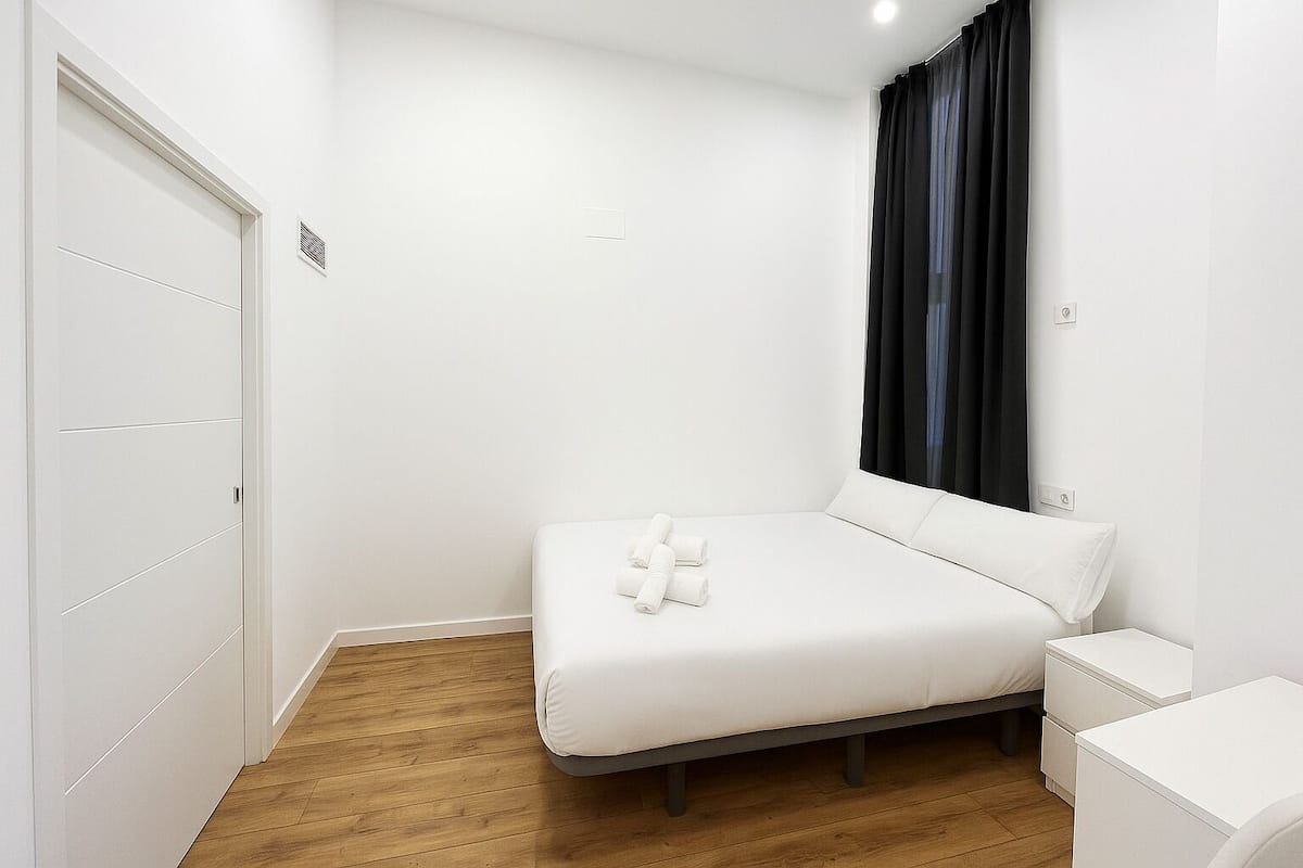 Un dormitorio con cama, mesita de noche, armario y ventana con cortinas.
