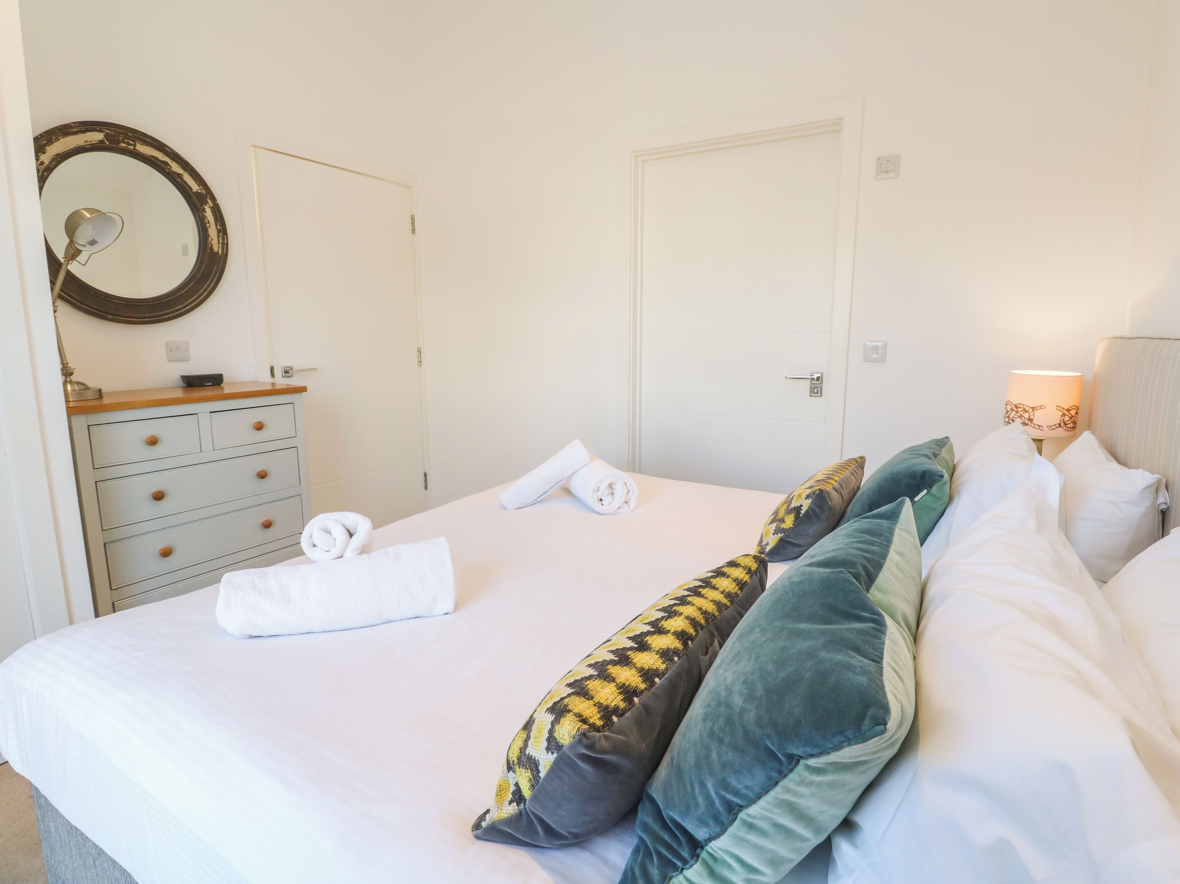 Een opgemaakt bed met witte lakens en opgerolde handdoeken, een dressoir met lades, een spiegel en een lamp op het nachtkastje.