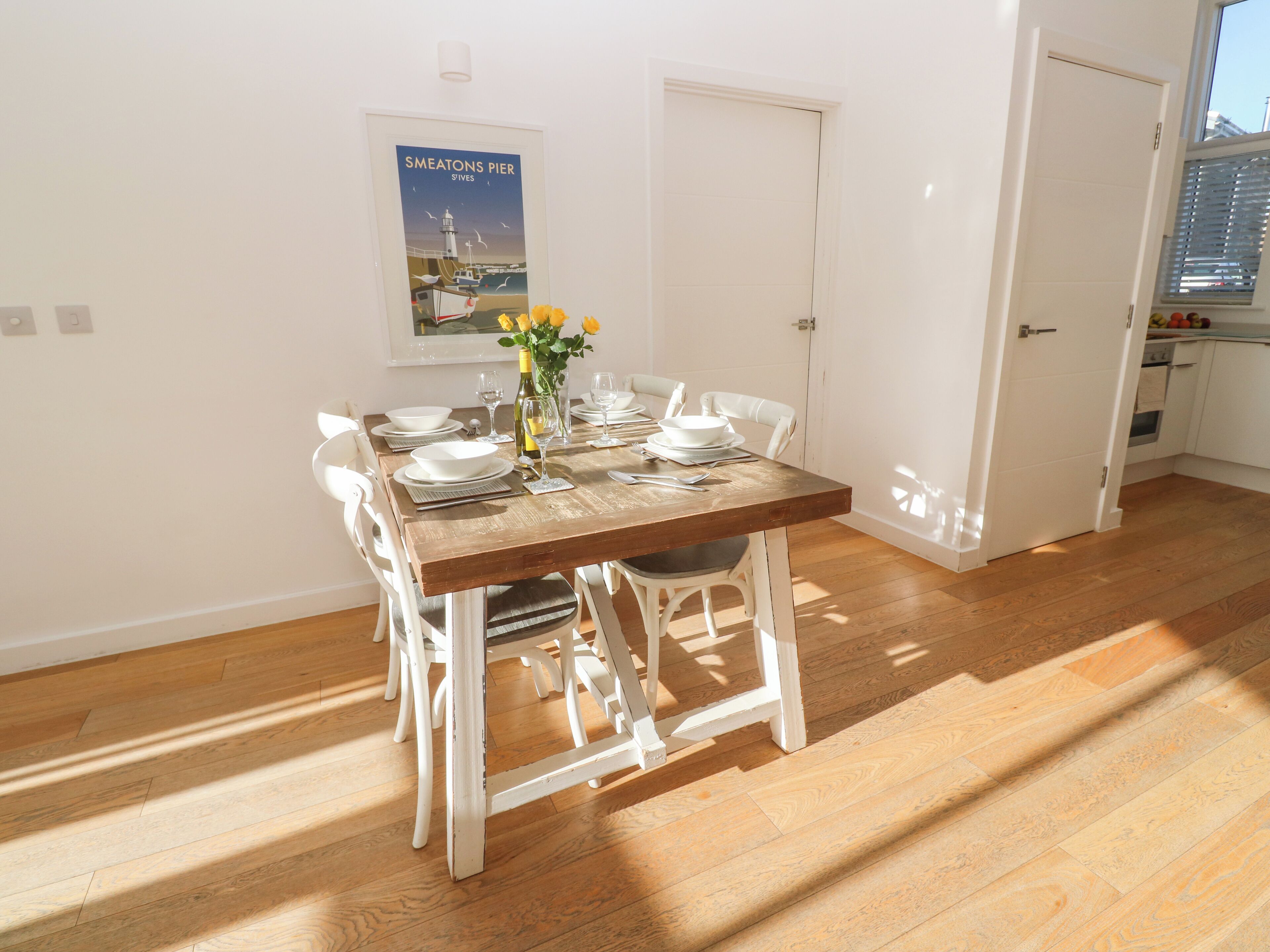 A dining area with a wooden table set for four, white chairs, and a poster on the wall.