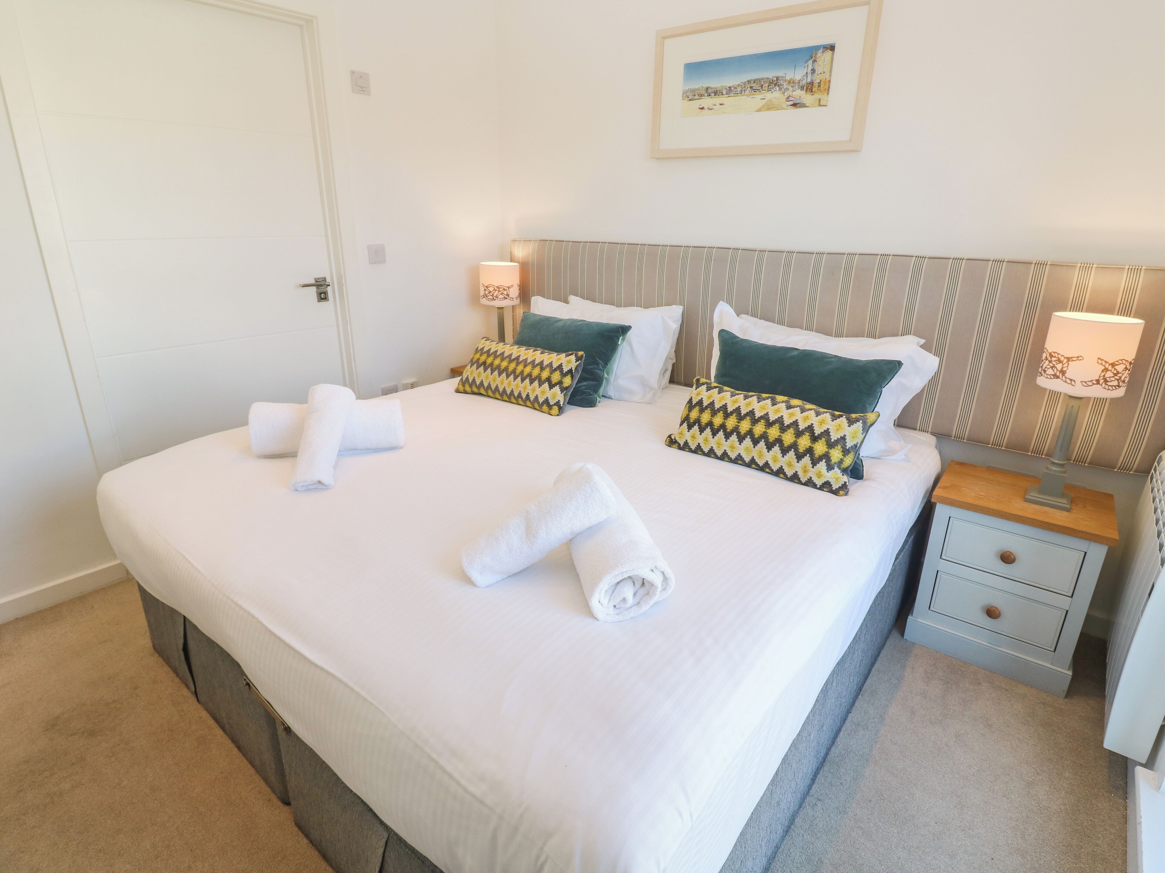 A neatly made bed with pillows and rolled towels, a bedside table with a lamp, and a framed picture on the wall.