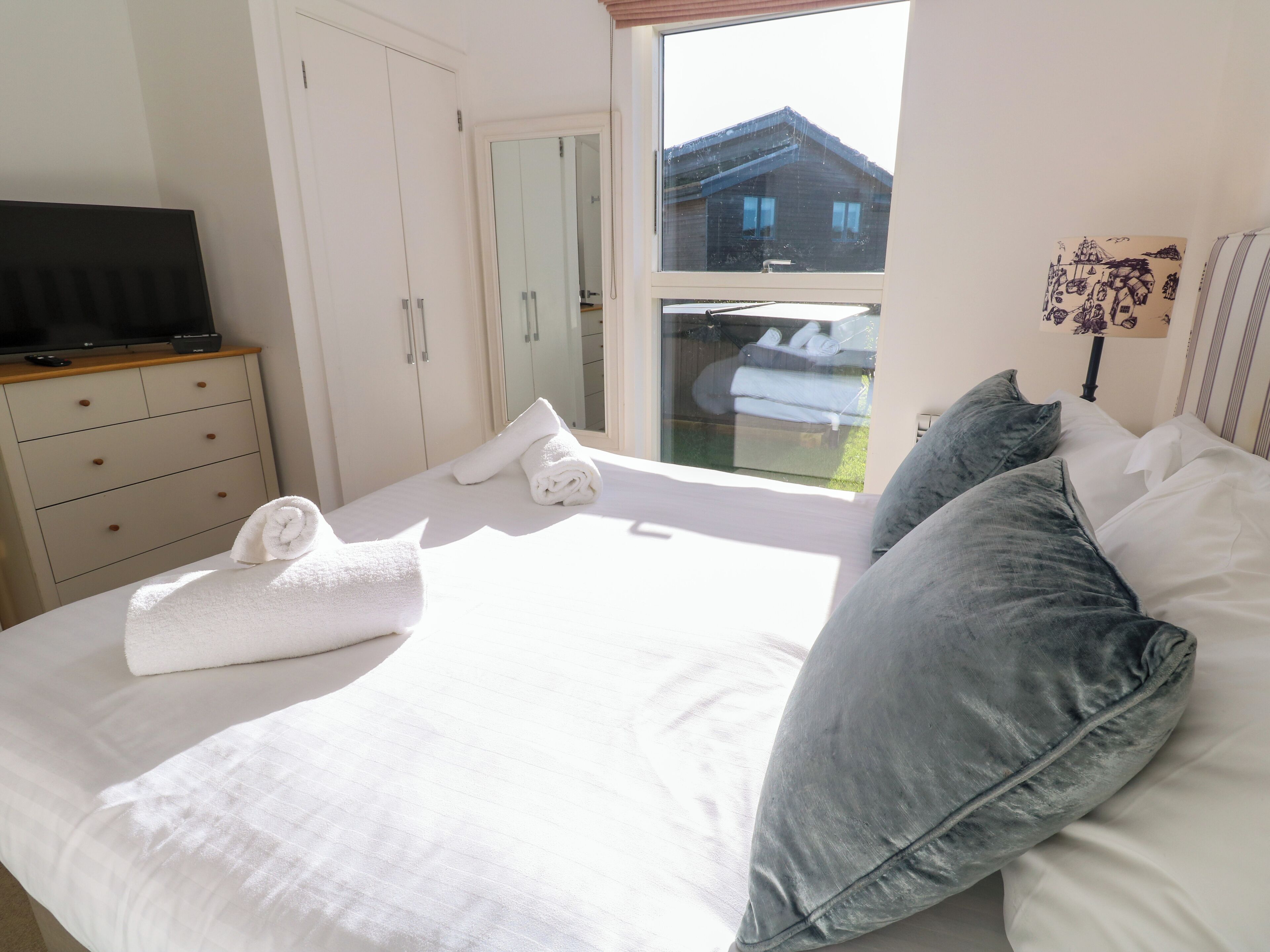 Un lit bien fait, avec du linge de lit blanc et deux serviettes enroulées, une commode en bois sur laquelle est posée une télévision, et une fenêtre donnant sur une maison et de la verdure.