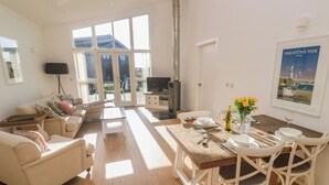 A spacious living room with a sofa, a dining table set for four, and a large window providing natural light. - Una, Lodge 77 (St Ives)