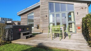 A modern building with a large glass door, a wooden deck with outdoor furniture, and a hot tub. - Una, Lodge 77 (St Ives)