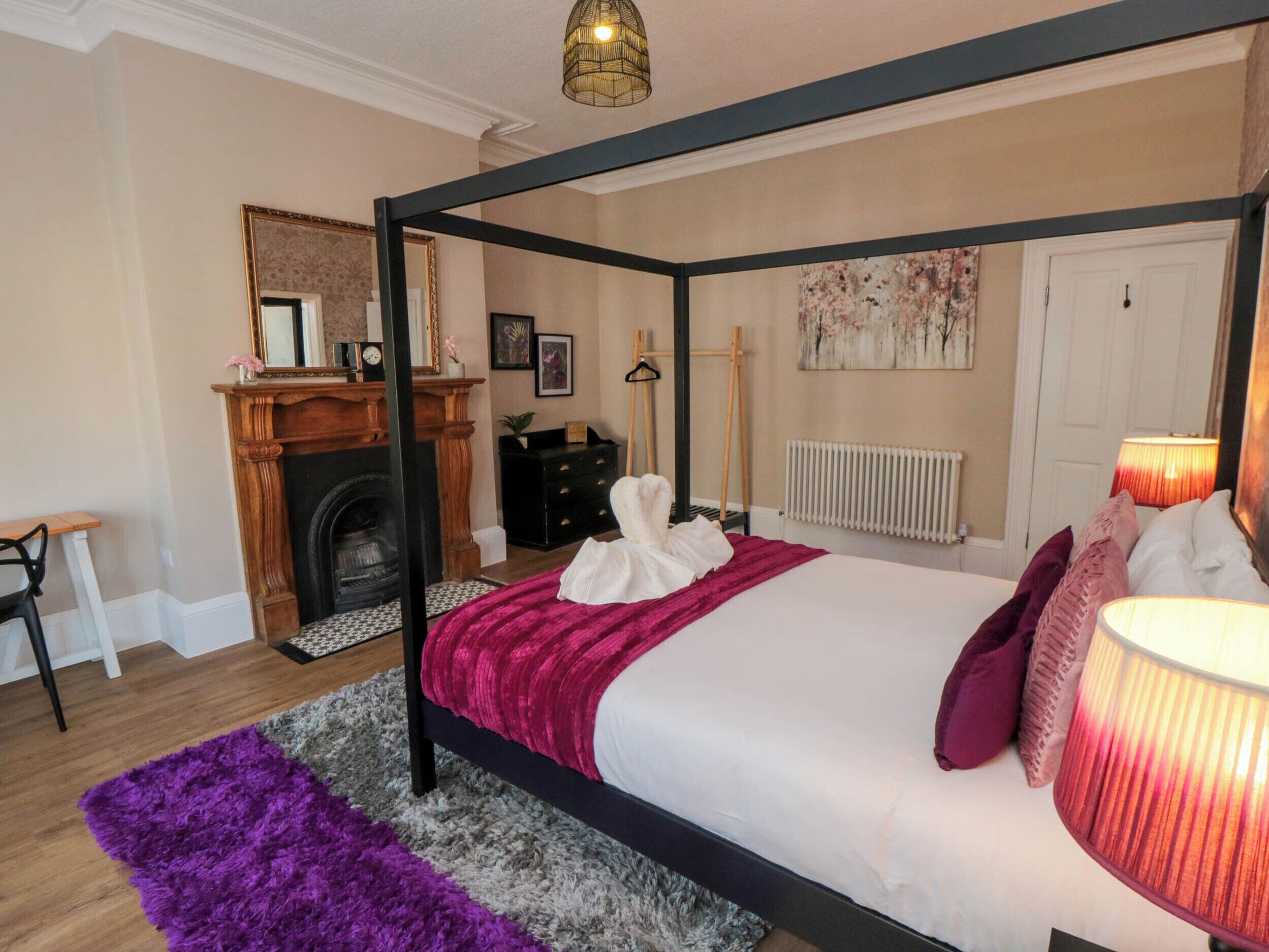 A four-poster bed with a white and purple bedspread, a black dresser, a fireplace, and a radiator.