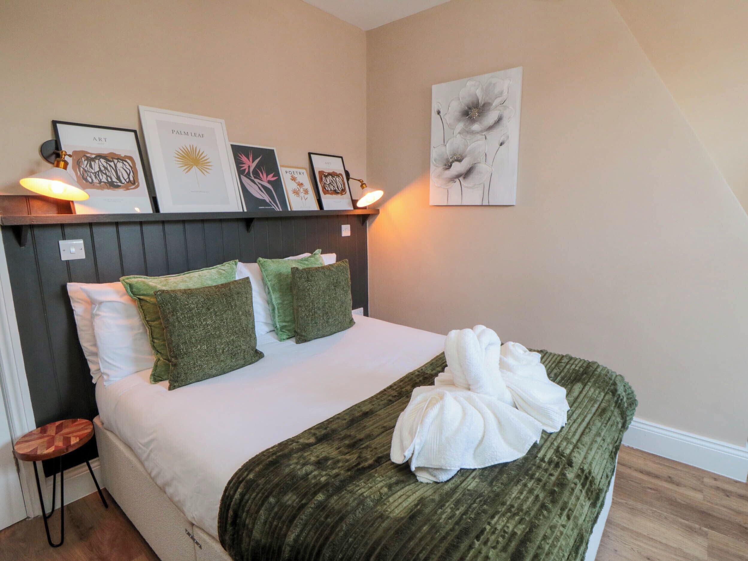 A neatly made bed with green and white pillows, a green blanket, and a towel on the bed. There are framed artworks on the wall above the bed.