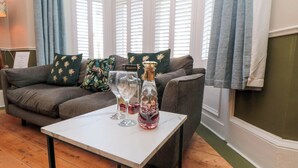 A cozy living room with a grey sofa, patterned cushions, a small white table with wine glasses and a decanter, and a window with white shutters. - Coronation House (Scarborough)