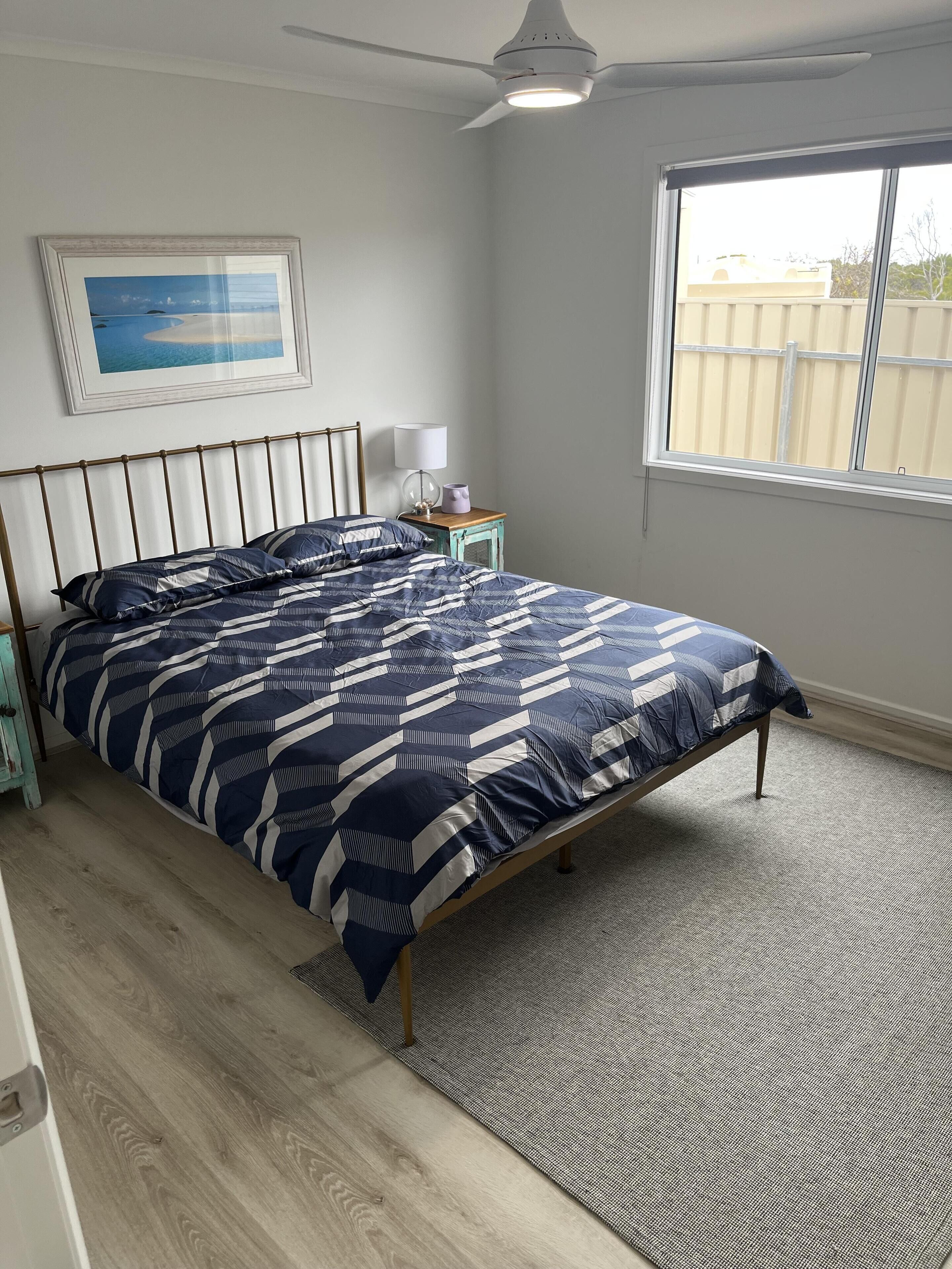 Un dormitorio con una cama, una mesita de noche, una lámpara, un ventilador de techo, una ventana y un cuadro en la pared.