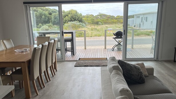 A modern living room with a wooden dining table, beige chairs, a large sofa, and a sliding glass door leading to an outdoor deck. - Clearwaters - Single story, 4-bedroom holiday home (Marion Bay)