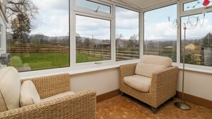 A room with a large bay window, wicker furniture, and a view of a rural landscape. - 14 Birchwood Road (Lydney)