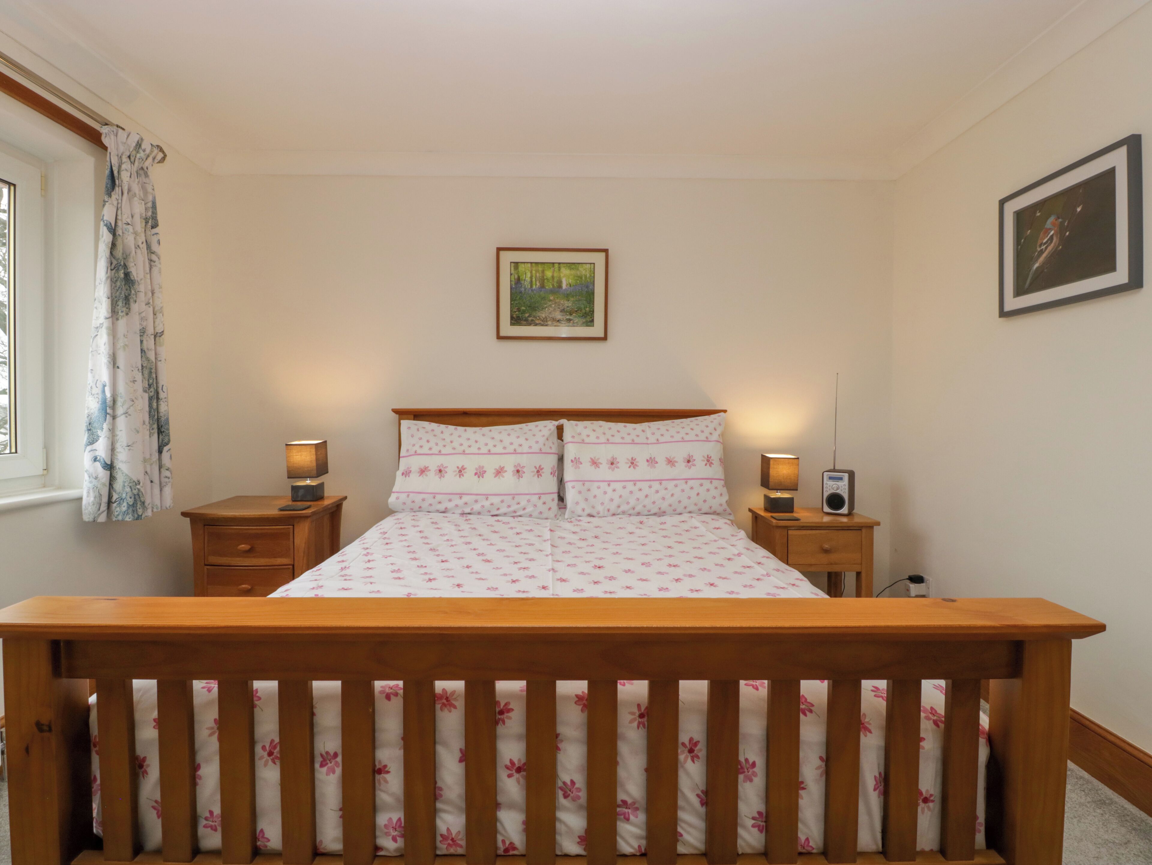 A bedroom with a wooden bed frame, a nightstand with a lamp, a radio, and a window with curtains.