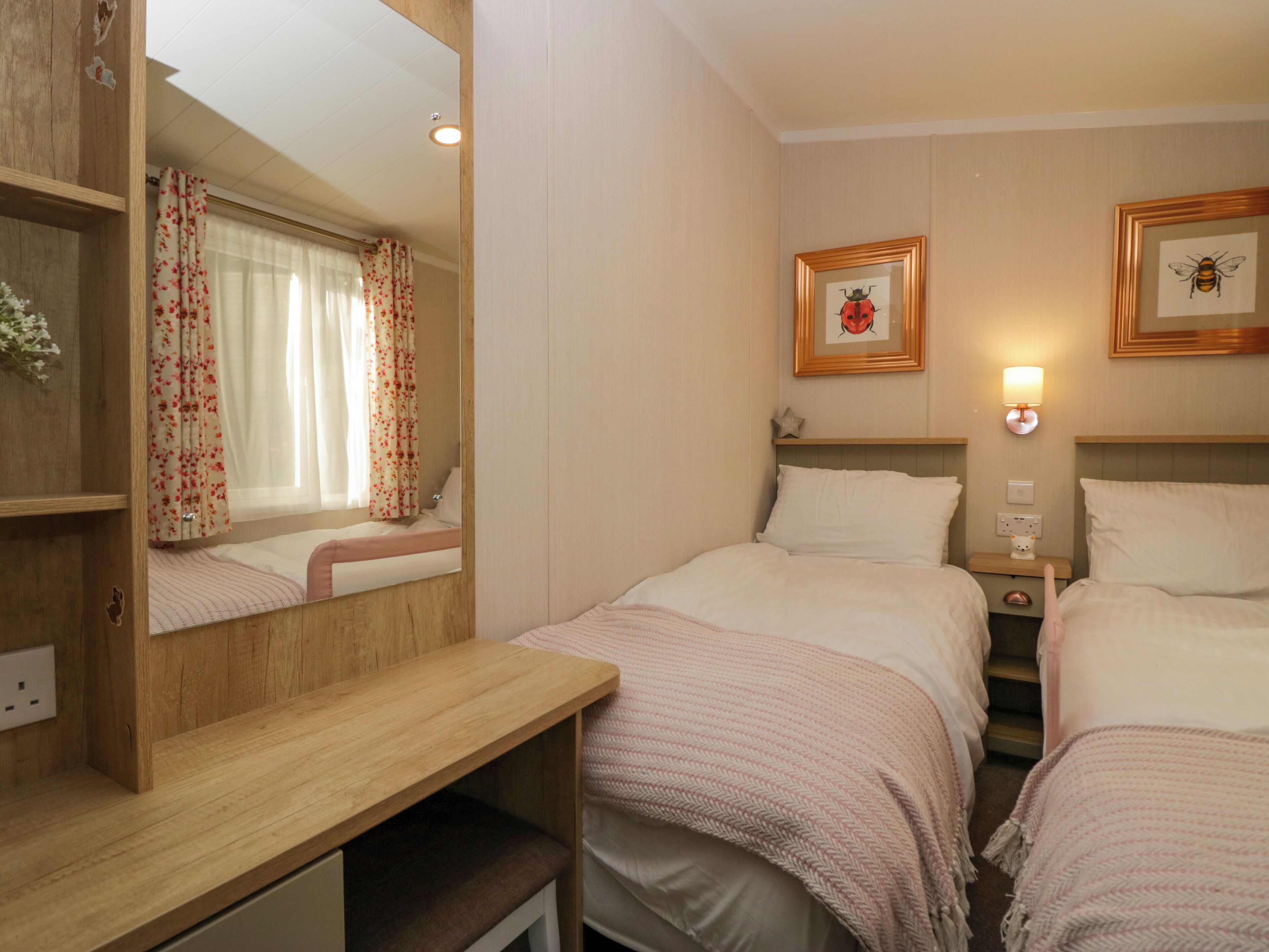 A hotel room with two beds, a wooden desk, a mirror, a window with floral curtains, and wall-mounted artwork.
