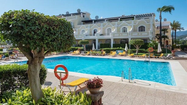 A pool area with lounge chairs and a large building in the background. - Serinamar - Aloha Gardens (Marbella)
