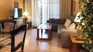 A living room with a sofa, a TV, a table, and a chair. - Serinamar - Aloha Gardens (Marbella)