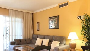 A living room with a grey sofa, a glass coffee table, a wooden chair, a lamp, and a potted plant. - Serinamar - Aloha Gardens (Marbella)