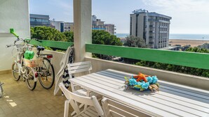 A balcony with a white table and chairs, a bicycle, and a view of the sea and hotel buildings. - Nice apartment with sunny terrace in Bibione (Bibione)