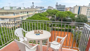 Un balcón con sillas y una mesa de plástico blanco, un letrero que dice "BEACH", y vistas de edificios y árboles.