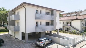 A modern two-story apartment building with balconies, a car parked in the driveway, and a neighboring building with a red-tiled roof. - Elegant and luminous apartment with terrace (Bibione)
