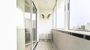 Ein schmaler Balkon mit gemusterten Bodenfliesen, zwei weißen Hockstühlen und hängenden Pflanzen.