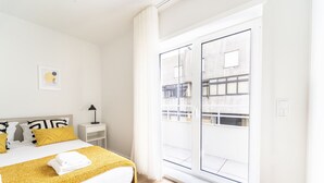 A hotel room with a bed, a nightstand, a lamp, a window with a view of buildings, and a yellow throw blanket. - Forum Apartments II by House and People (Braga)
