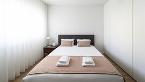 Ein modernes Hotelzimmer mit einem großen Bett, zwei Kissen, einem Nachttisch mit Lampe und einem Fenster mit durchsichtigen Vorhängen.