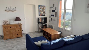 A living room with a blue sofa, wooden dresser, TV, and a window with a view. - Grain de Café Barneville Plage (Barneville-Carteret)