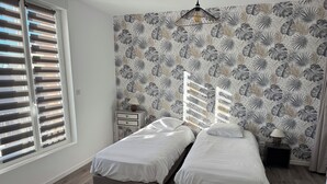 A bedroom with two single beds, a nightstand, a lamp, and a window with blinds. - Grain de Café Barneville Plage (Barneville-Carteret)