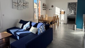 A modern living room with a blue sofa, wooden dining table, and chairs. There is a window with a view of a brick building outside. - Grain de Café Barneville Plage (Barneville-Carteret)
