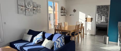 A modern living room with a blue sofa, wooden dining table, and chairs. There is a window with a view of a brick building outside.