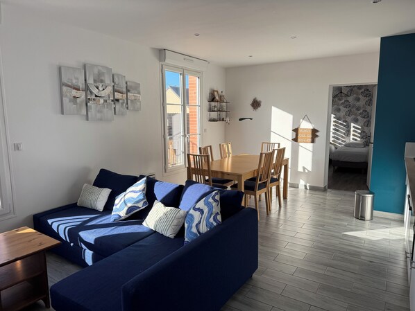 A modern living room with a blue sofa, wooden dining table, and chairs. There is a window with a view of a brick building outside.