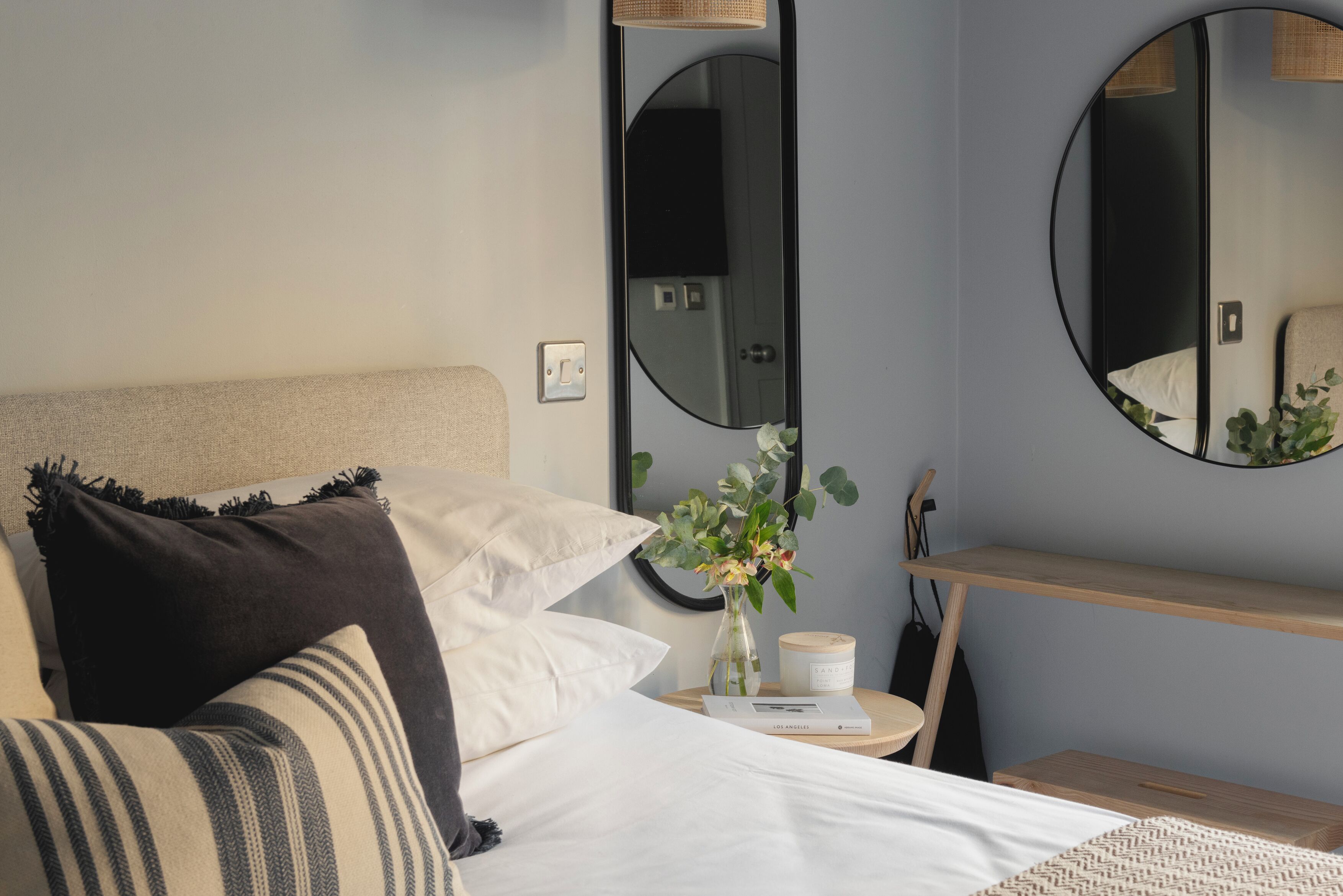 Una camera da letto ordinata con un letto, cuscini, un comodino con un vaso e una candela, e due specchi appesi al muro.