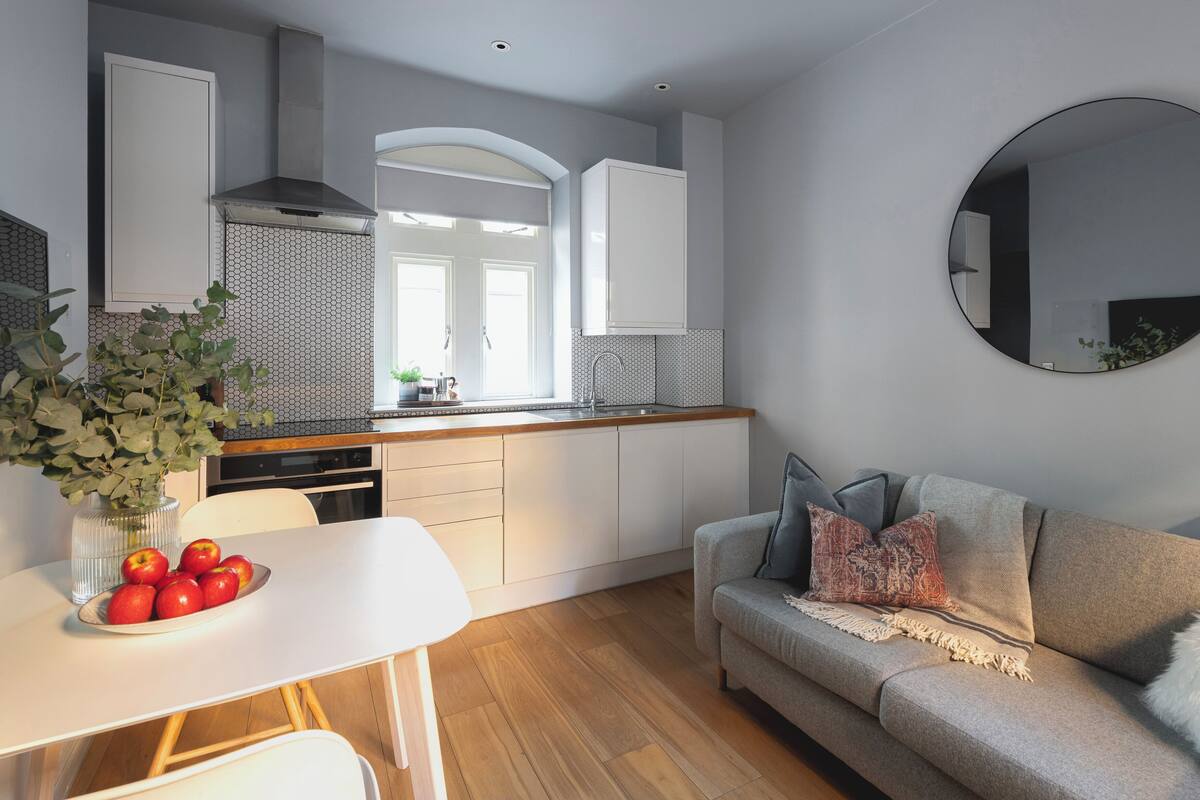 A modern kitchen with white cabinets, a dining table with apples, and a grey sofa.