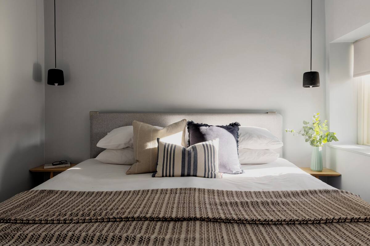 A neatly made bed with a striped blanket, a headboard, and multiple pillows. There are two bedside tables with lamps, and a vase with flowers on the right side.