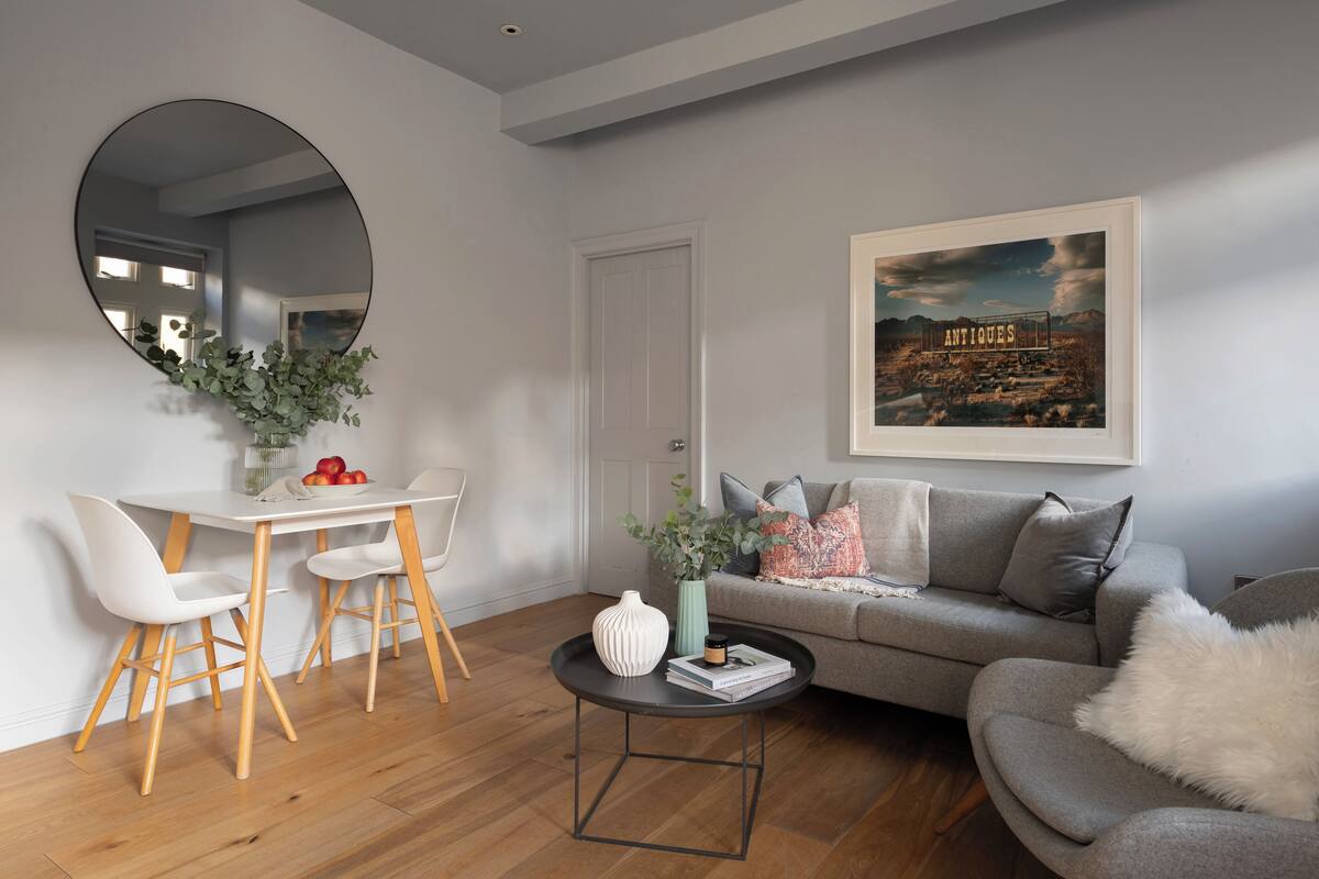 A modern living room with a round mirror, a dining table with chairs, a grey sofa, and a framed picture on the wall.