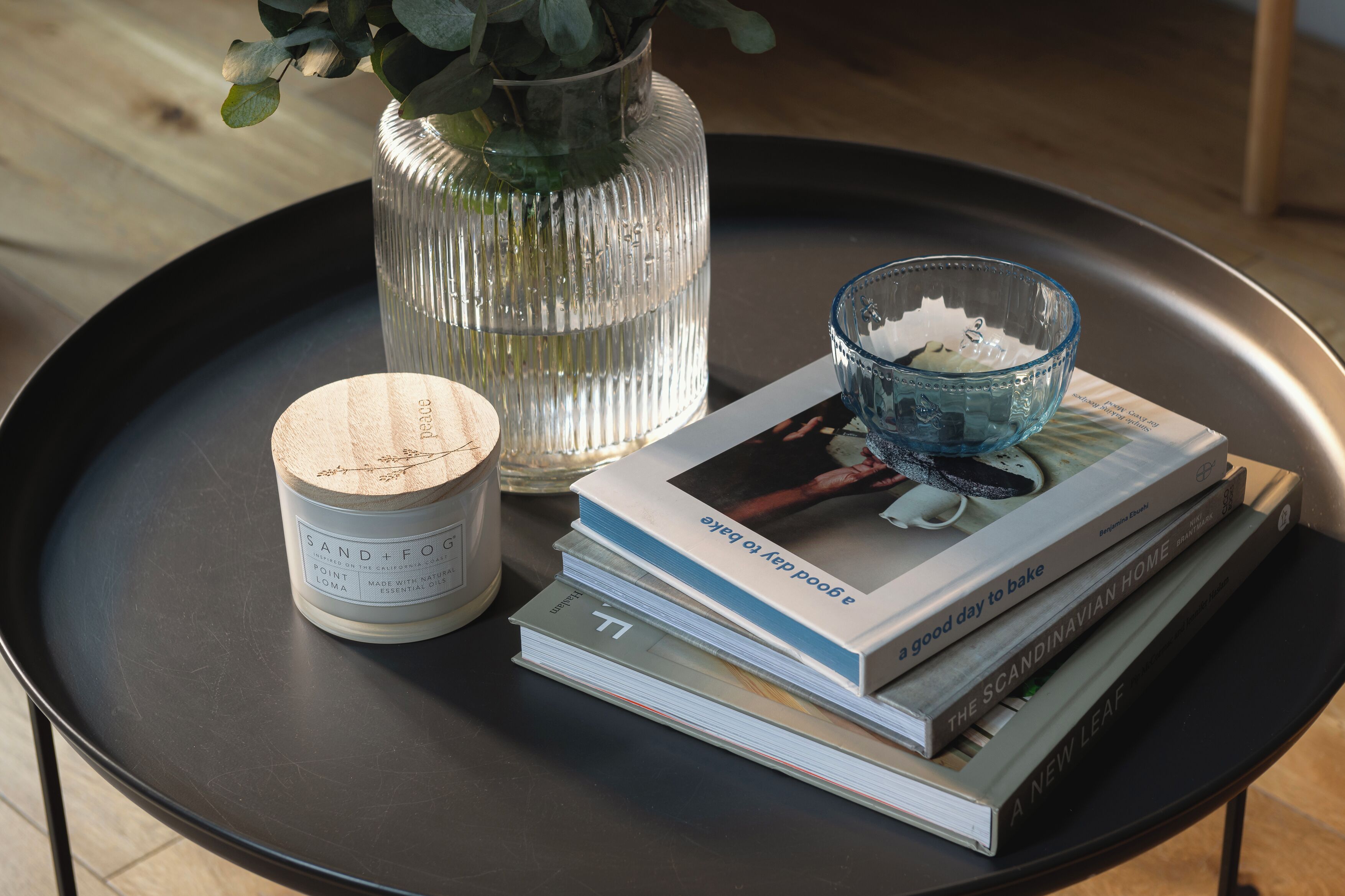 A black tray with a candle, a glass vase with green leaves, and a stack of books on a wooden floor.