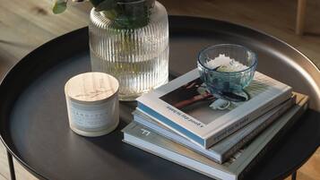 A black tray with a candle, a glass vase with green leaves, and a stack of books on a wooden floor.